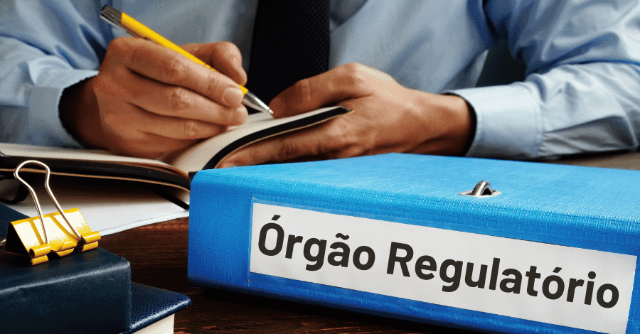 A imagem mostra uma pessoa de terno escrevendo em um caderno com um lápis, enquanto na frente dela, sobre a mesa, há uma pasta azul com o rótulo "Órgão Regulatório". A pasta está em destaque, sugerindo que o conteúdo está relacionado a regulamentações ou fiscalização. Ao lado da pasta, há outros itens de escritório, como um clipe de papel grande e outra pasta parcialmente visível.